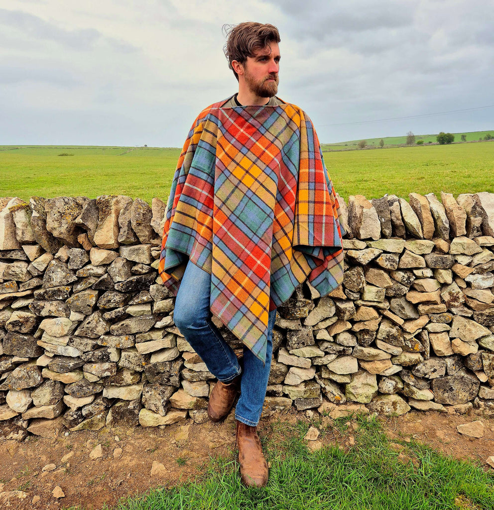 A man wearing a colorful plaid wool poncho, standing by a stone wall in a scenic countryside setting.
