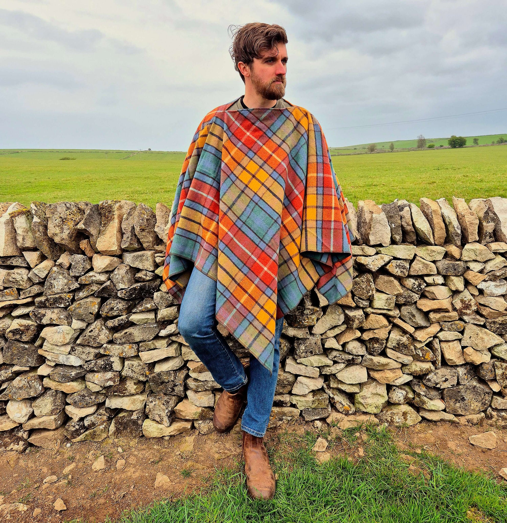 A man wearing a colorful plaid wool poncho, standing by a stone wall in a scenic countryside setting.