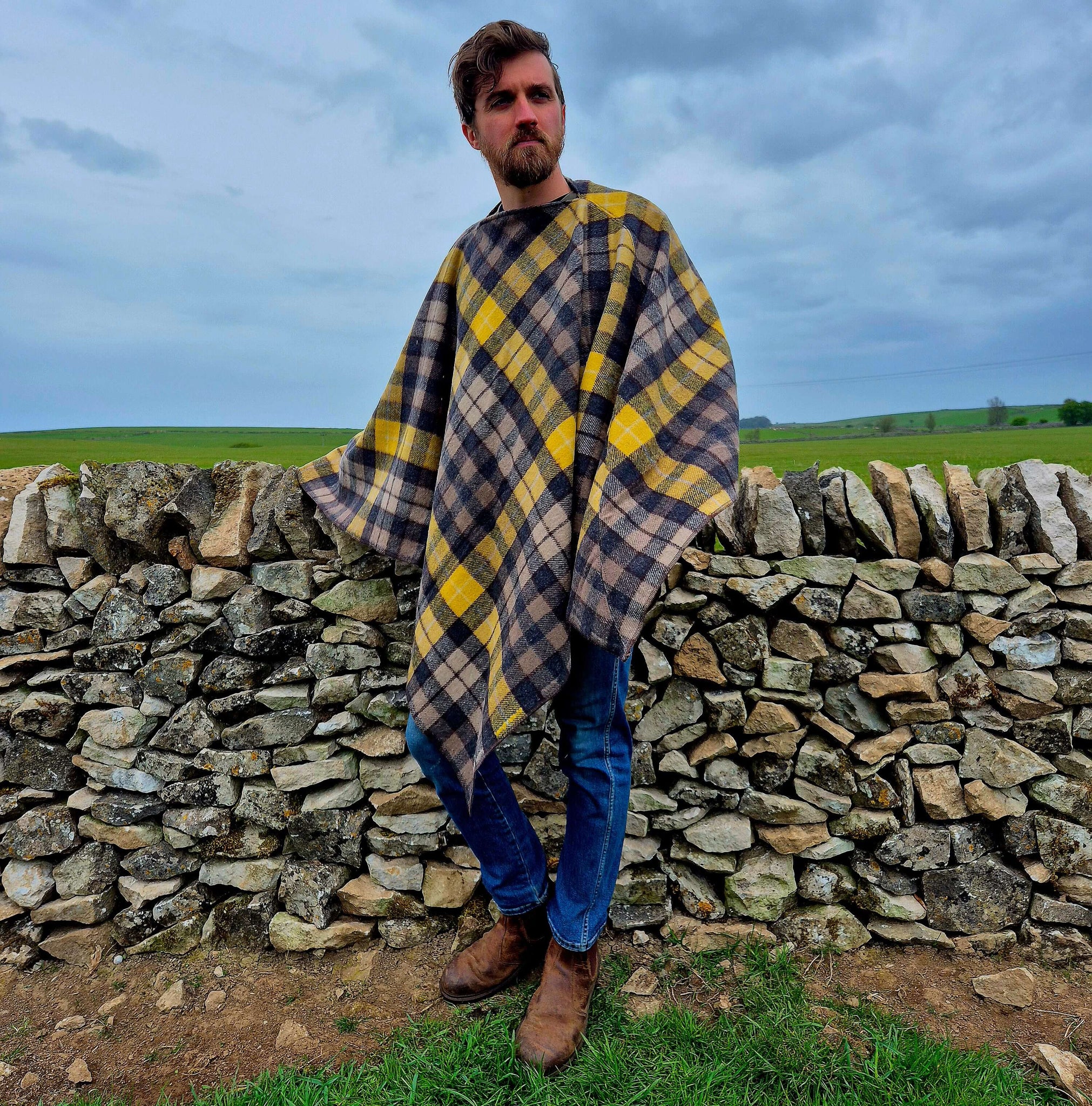 A man wearing a plaid wool poncho standing by a stone wall, showcasing warmth and style in an outdoor setting.