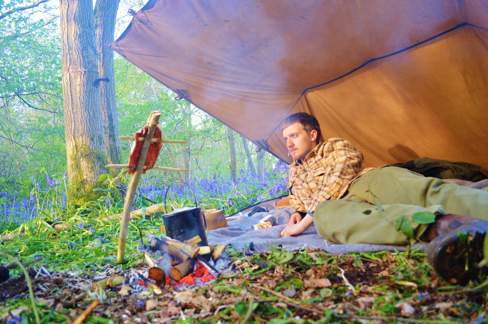 A person relaxing under a brown wax canvas tarp in a outdoor setting with trees and bluebells.