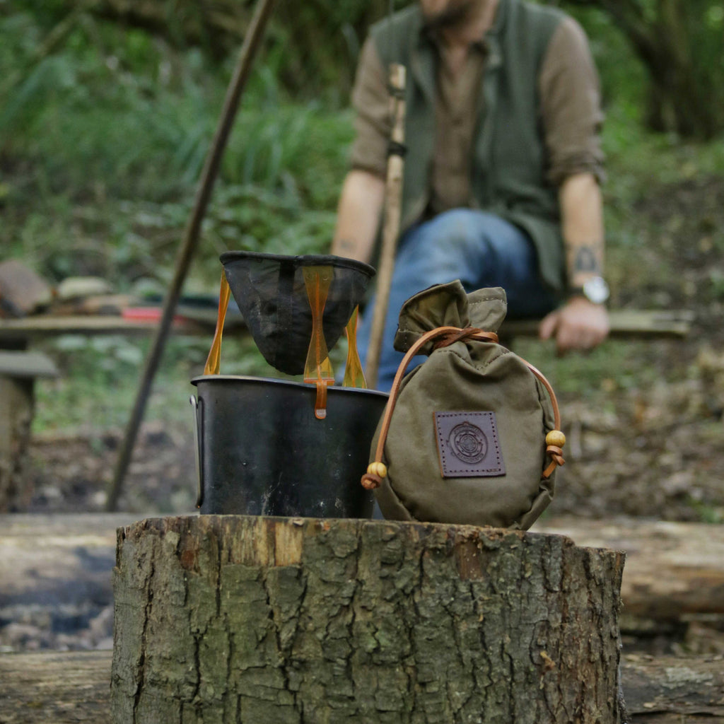 A cup of coffee on a tree stump with a wax canvas Sami style coffee pouch in the woods