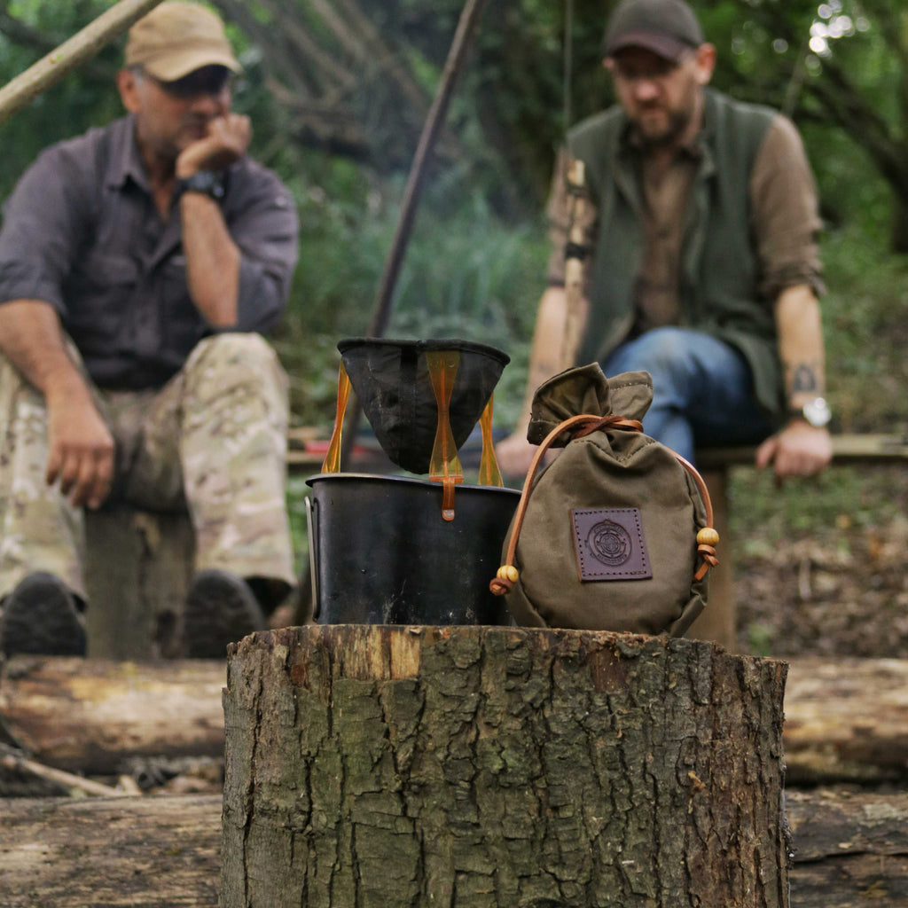 Men sit around the campfire watching their coffee brew with a wax canvas coffee pouch in the forefront